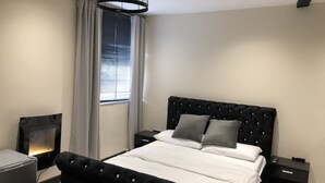 Een moderne slaapkamer met een zwart gestoffeerd bed, witte beddengoed, een zwart nachtkastje met een afstandsbediening, en een lamp aan de linkerkant.