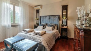 Een slaapkamer met een bed, nachtkastjes, een dressoir, een kroonluchter en een raam met gordijnen.