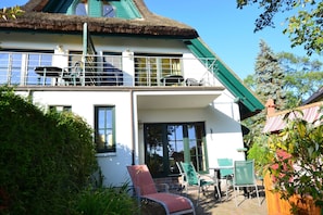 A two-story house with a thatched roof, a balcony with chairs, and a patio area with a table and chairs. - 17g balaclava 5 persons behind - Reethäuser 17 / Fam. Kröning / TZR (Gager)