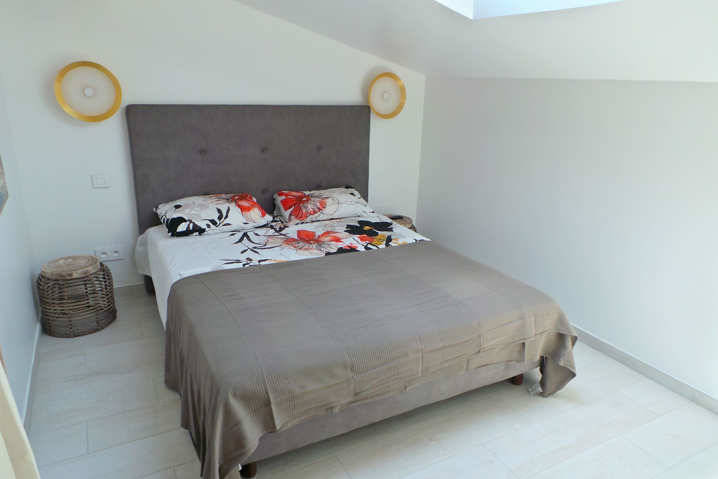 A bedroom with a bed, a grey headboard, a woven stool, and a wall-mounted light fixture.