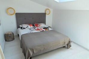 A bedroom with a bed, a grey headboard, a woven stool, and a wall-mounted light fixture.