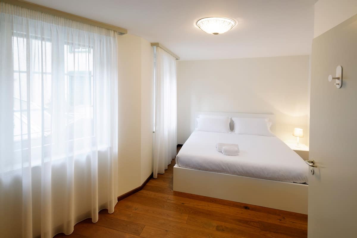 Un dormitorio con cama, mesita de noche, lámpara y ventana con cortinas traslúcidas.