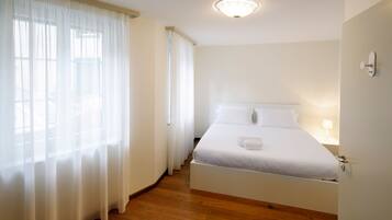 A bedroom with a bed, a nightstand, a lamp, and a window with sheer curtains.