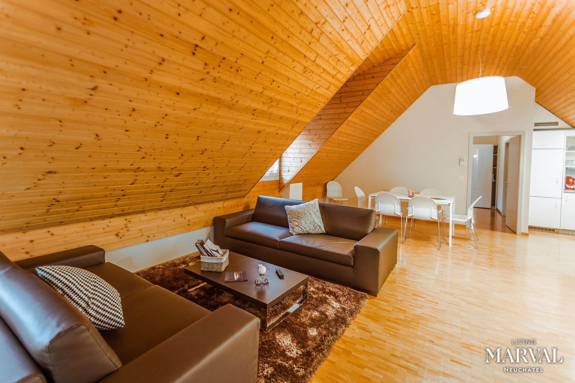 Una sala de estar moderna estilo loft con techo inclinado, un sofá de cuero marrón, una mesa de centro y un comedor con sillas blancas y una pequeña mesa.