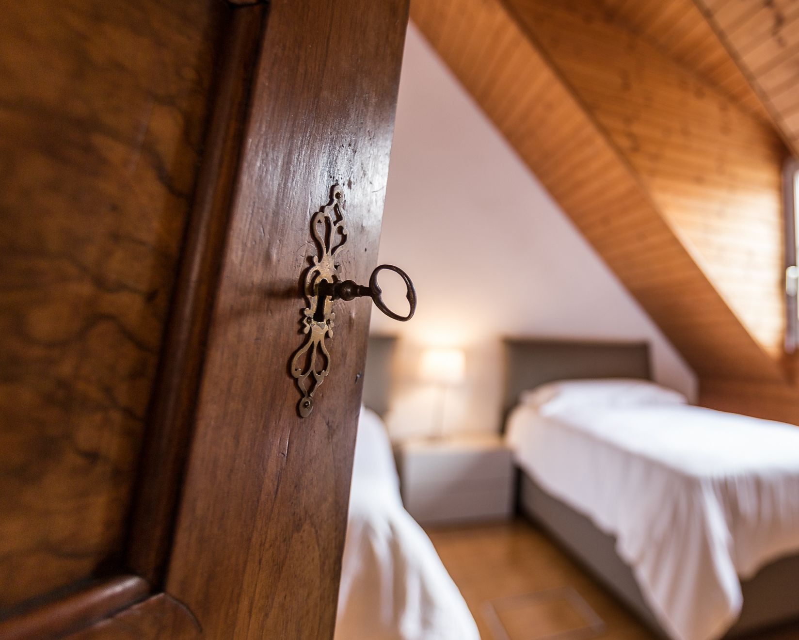 A wooden door with an ornate keyhole, a bed, and a nightstand in a room with wooden beams on the ceiling.