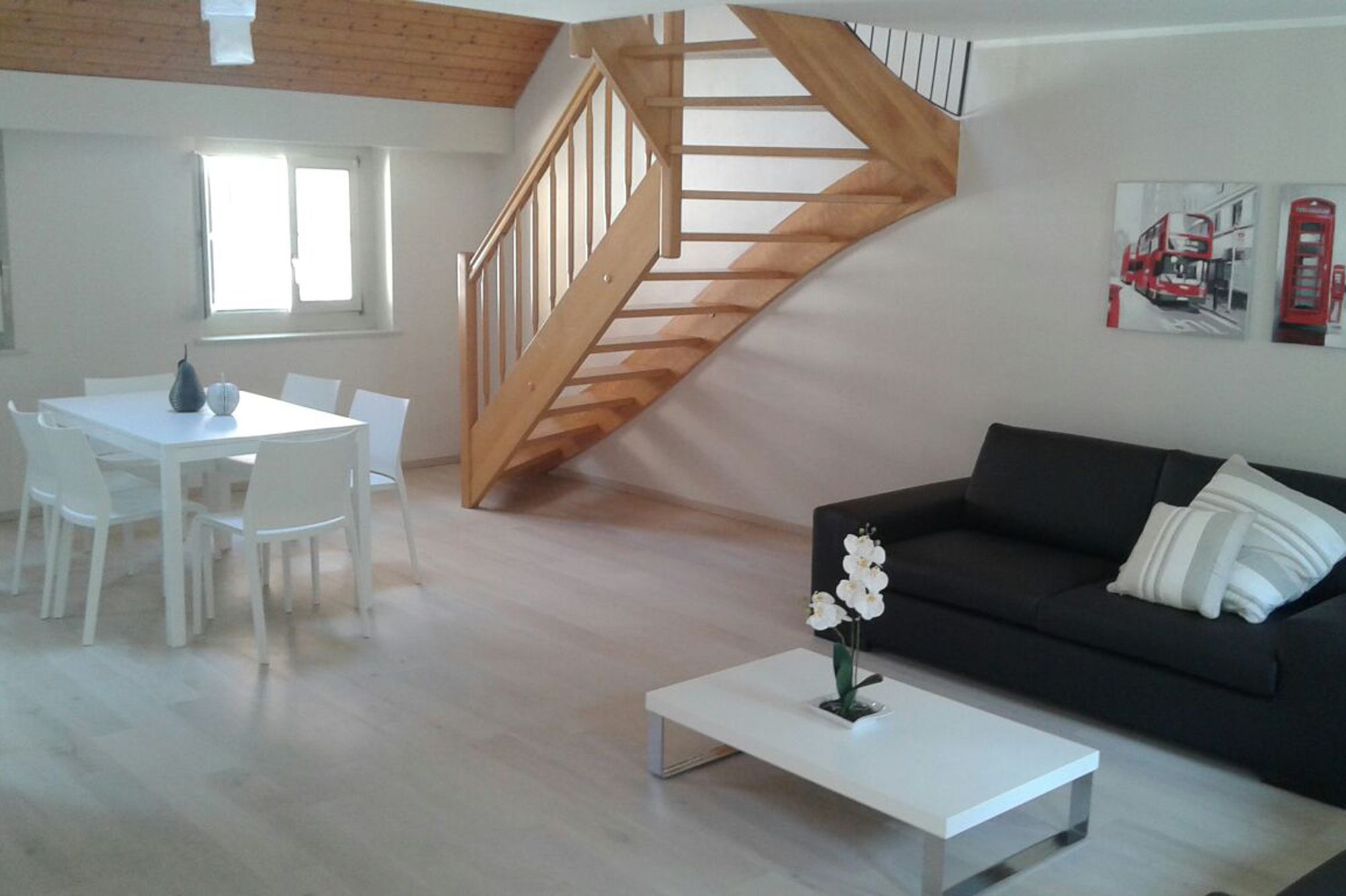 A modern living room with a staircase, a dining area with white chairs, and a black sofa with white cushions.