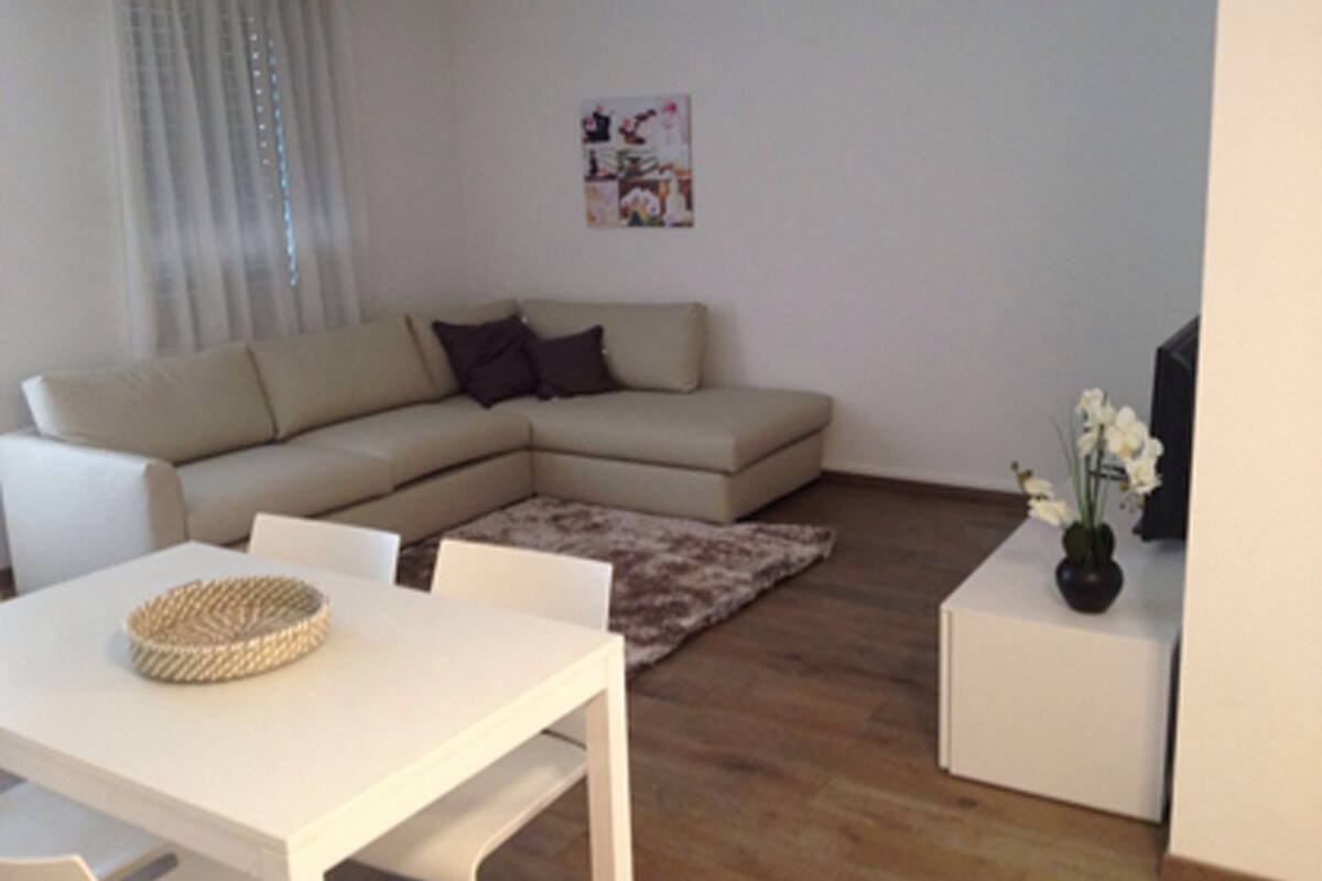 Una sala de estar moderna con un sofá seccional color beige, una mesa de comedor blanca y una pequeña mesita de noche blanca con un florero.