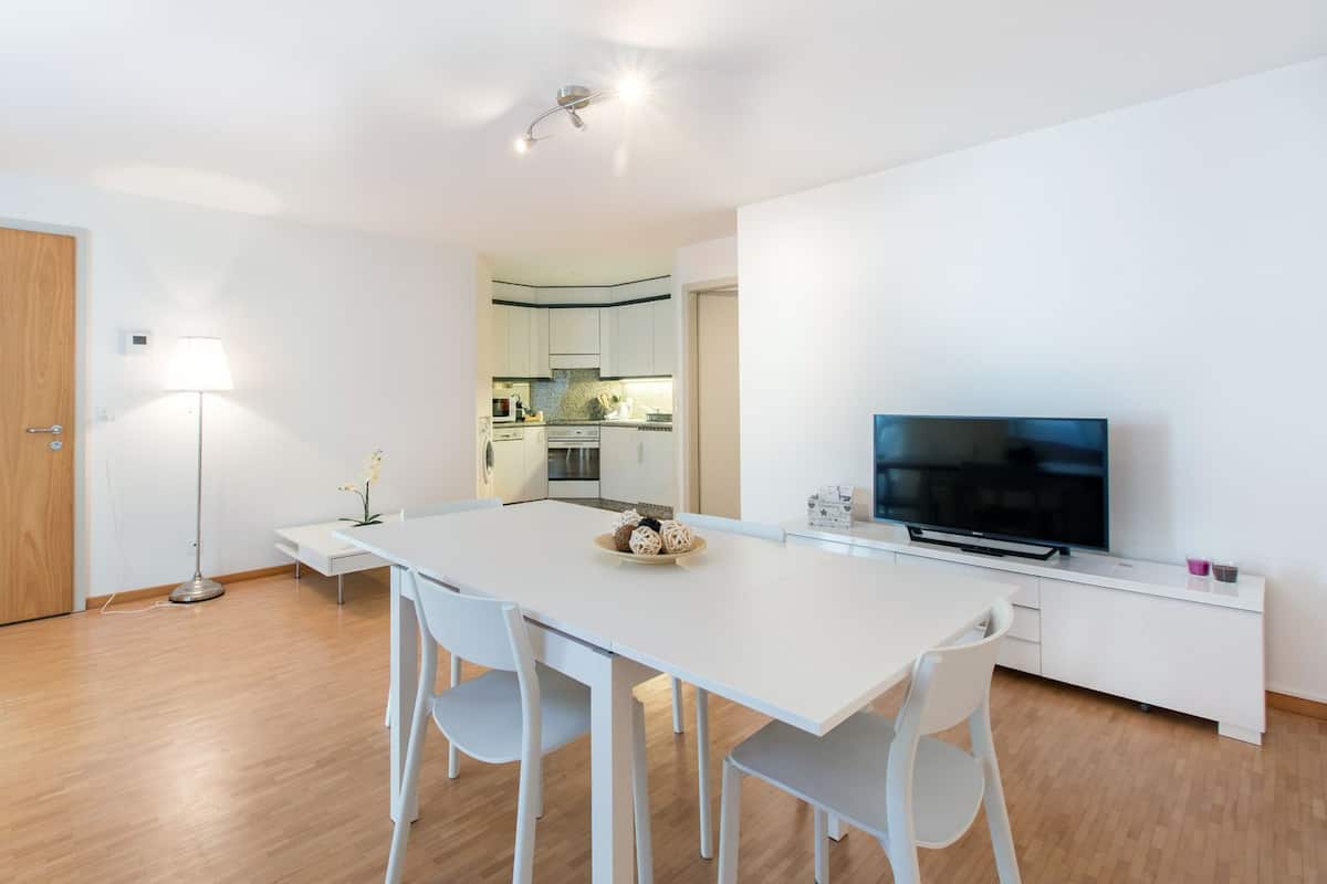 Una cocina moderna con armarios blancos, horno empotrado y comedor con mesa y sillas blancas.