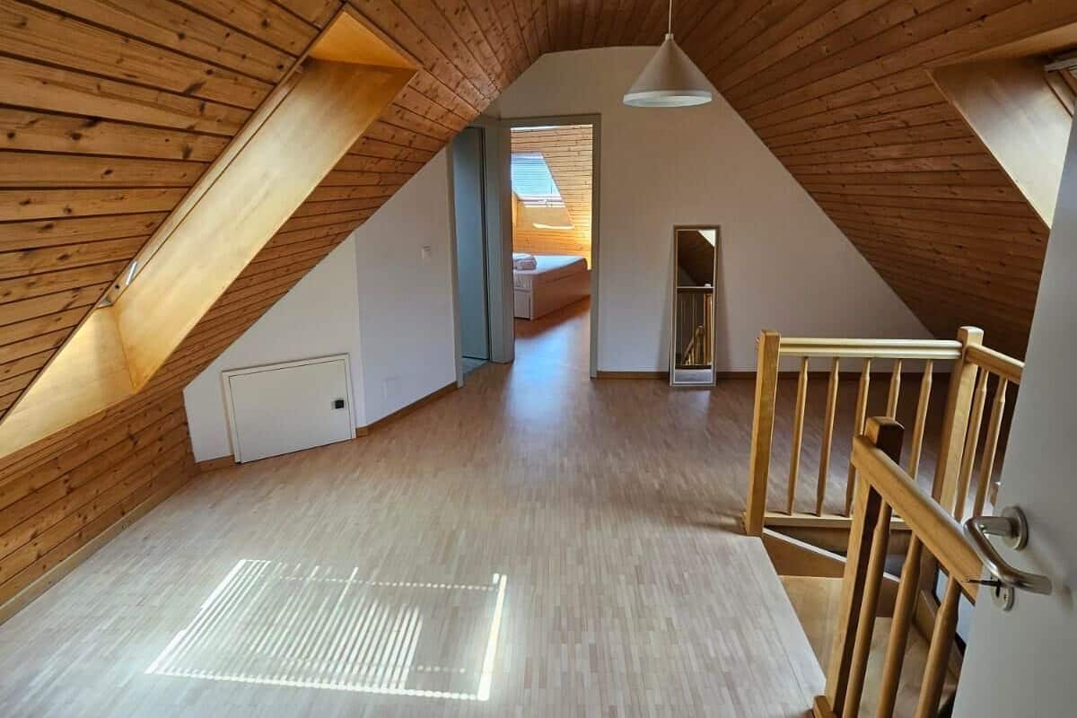 Un amplio ático con listones de madera en el techo, un tragaluz visible y vista a otra habitación.