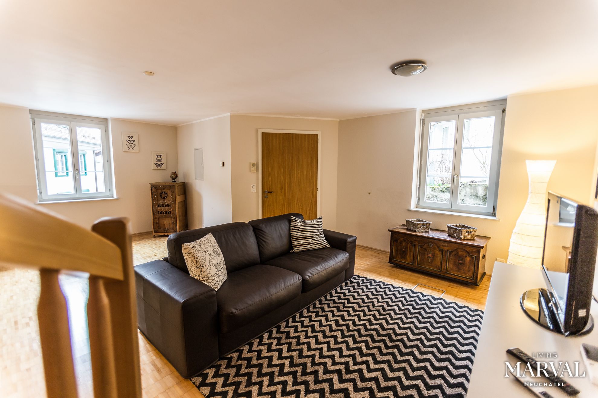 A living room with a black leather sofa, a patterned rug, wooden furniture, and large windows.