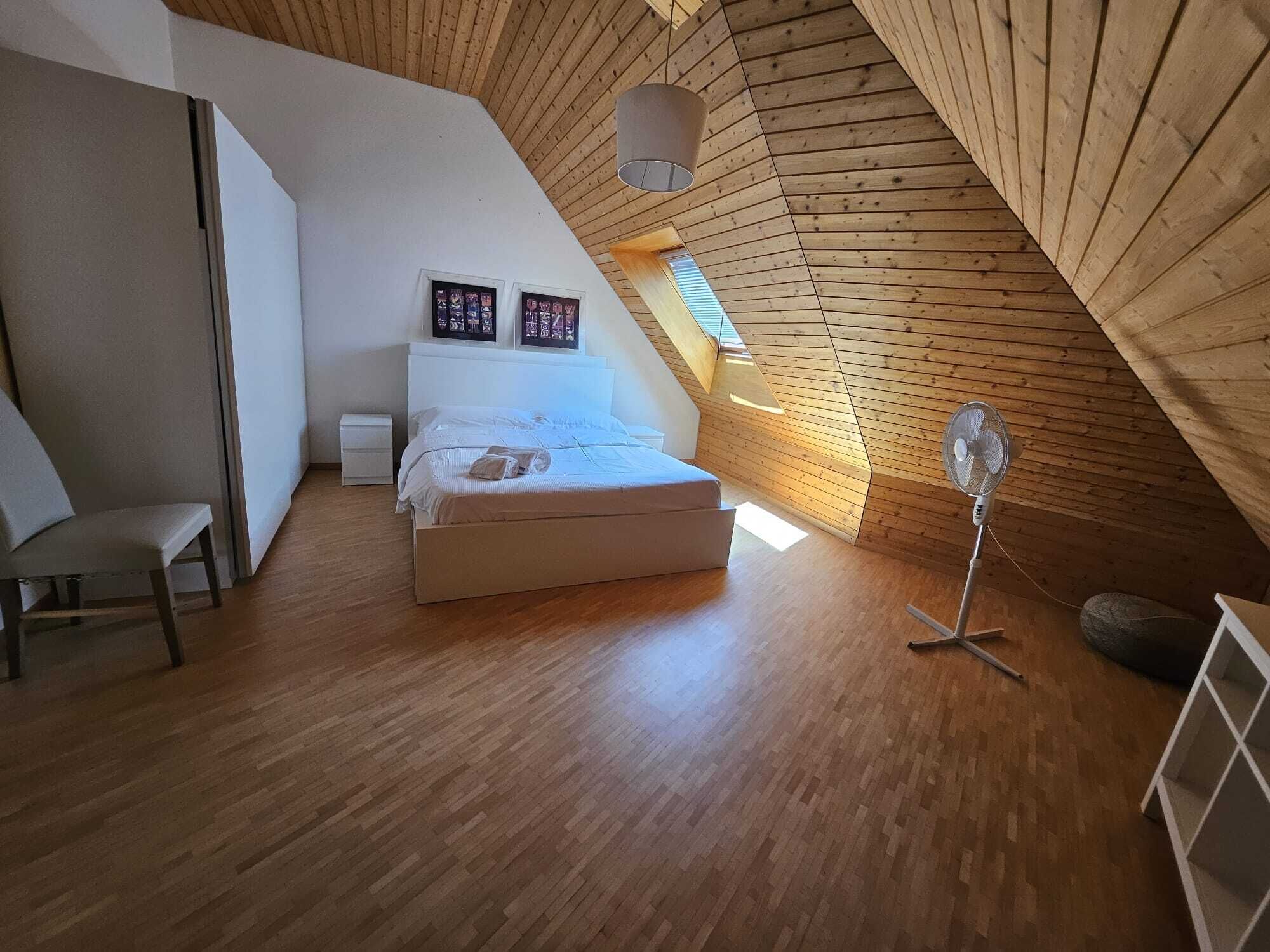 A bedroom with a sloped ceiling, wooden floor, a bed, a chair, a standing fan, and two framed pictures on the wall.