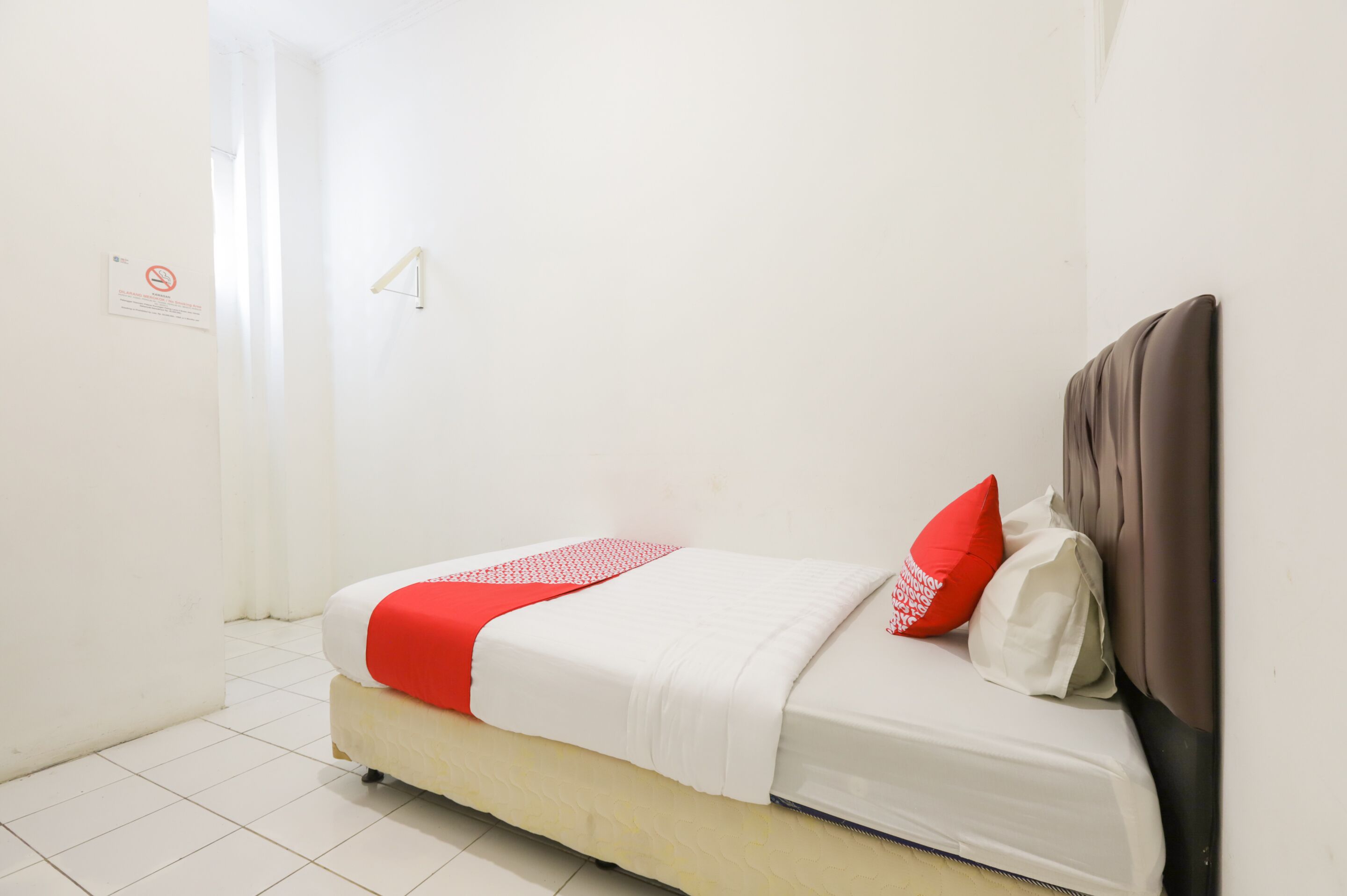 A single bed with a headboard, white bedding, and a red accent pillow.