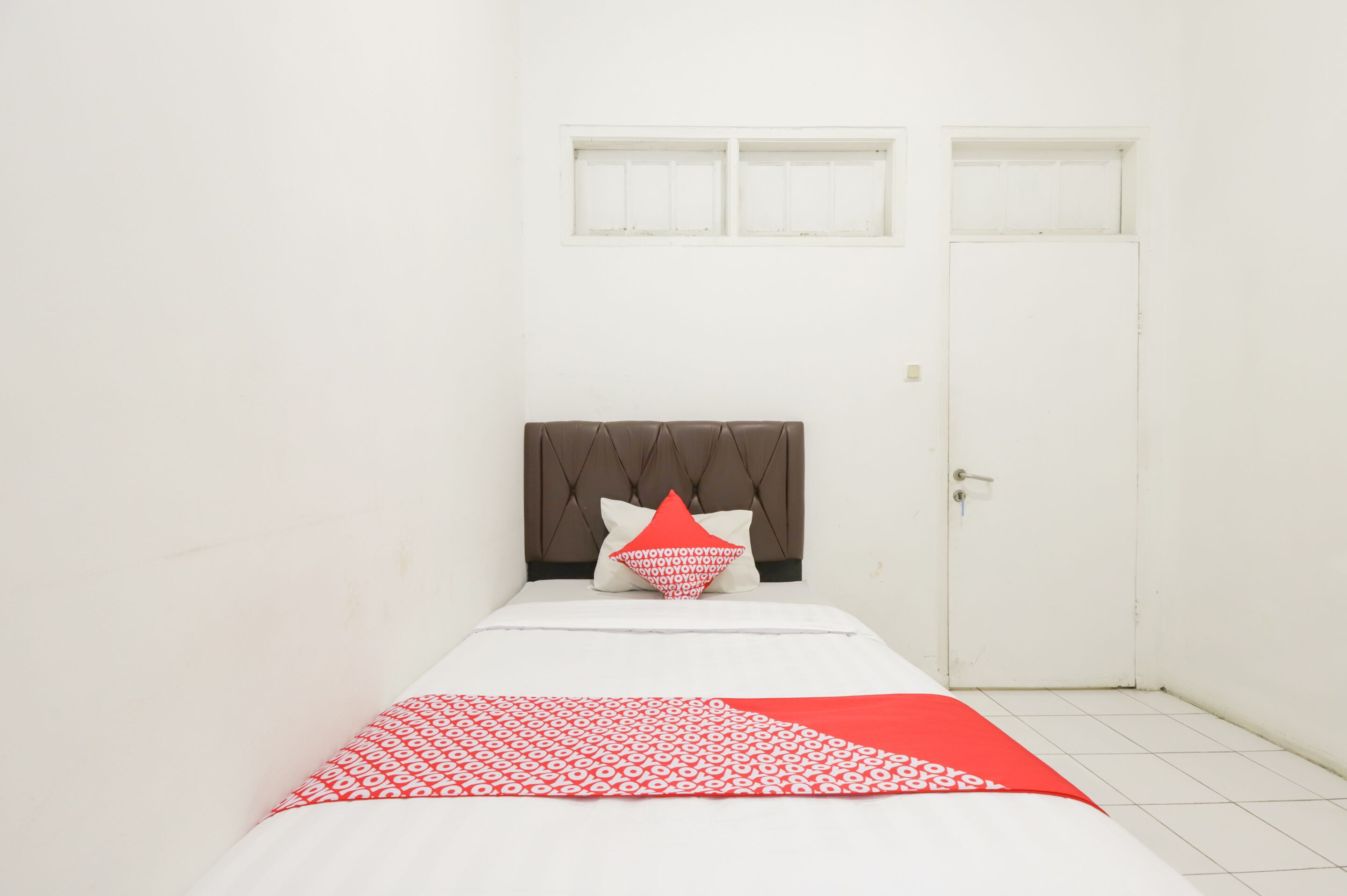 Een kleine, witte kamer met een enkel bed, een raam en een deur.