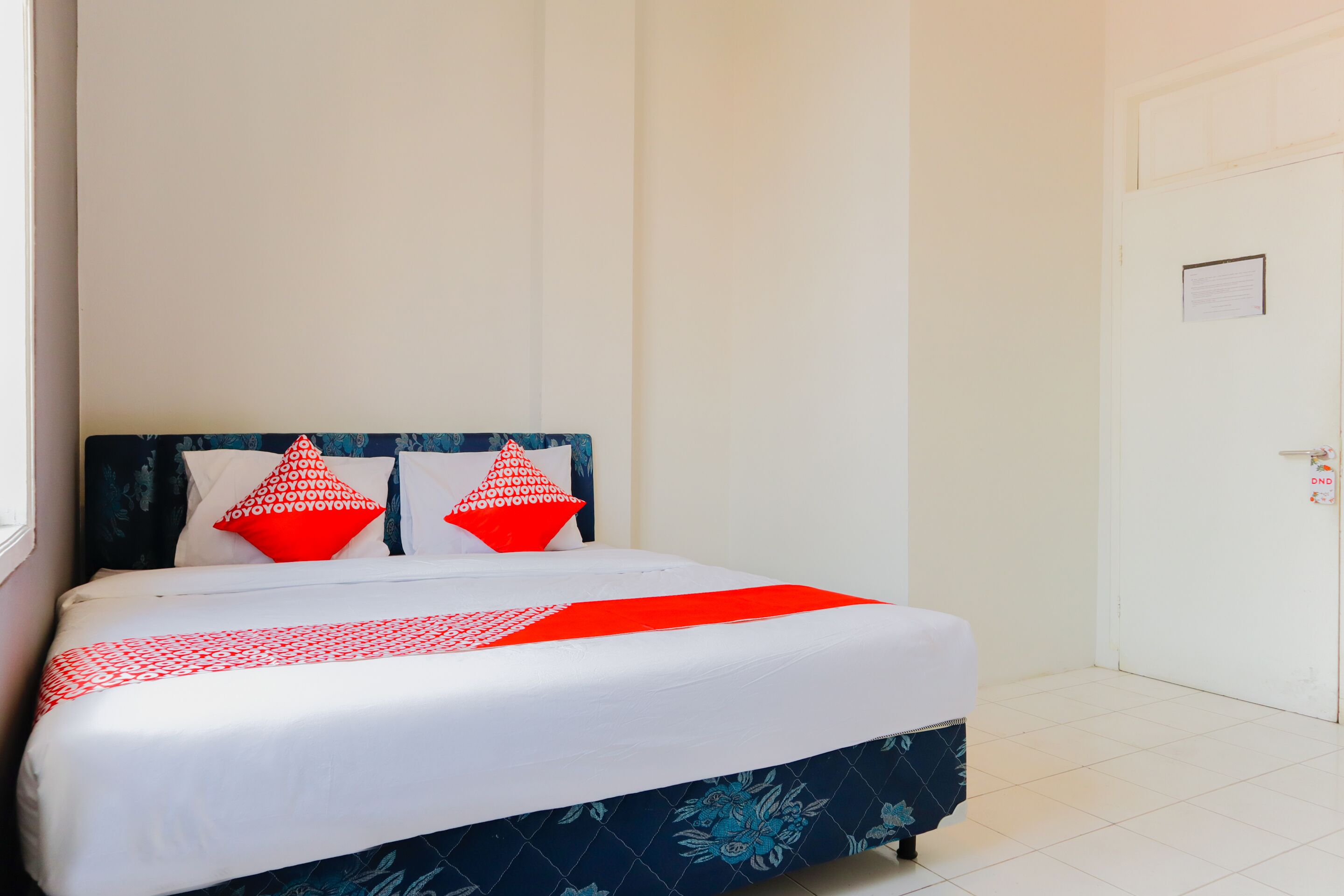 Un lit bien fait, recouvert d’une couvre-lit rouge et blanche, avec deux oreillers rouges et un couvre-lit bleu à motifs floraux.