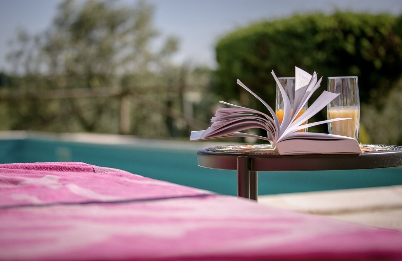 A poolside table with an open book, a glass of orange juice, and a glass of white beverage.