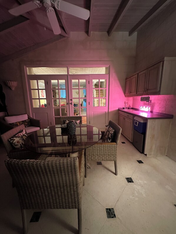 Een eetgedeelte met een tafel met glazen blad, omringd door rieten stoelen, een roze verlichte deur en een keuken met kasten en een vaatwasser.
