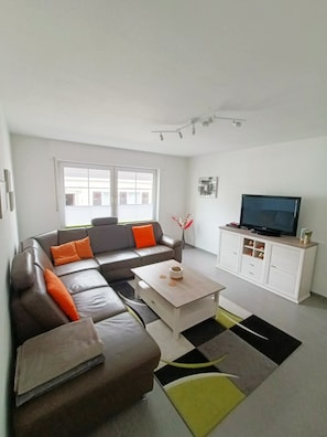 Ein modernes Wohnzimmer mit einem Ecksofa, einem Couchtisch, einem Fernseher auf einer Kommode und einem großen Fenster.