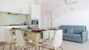 A modern kitchen with white cabinets, a dining table with chairs, and a blue sofa in a living area. - Salento Villa Emma - Green Apartment with Garden (Porto Cesareo)