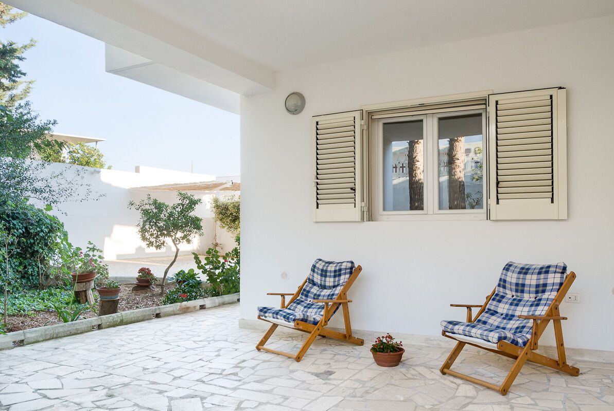 Un patio con dos sillones de madera, una pared blanca y una ventana con contraventanas.