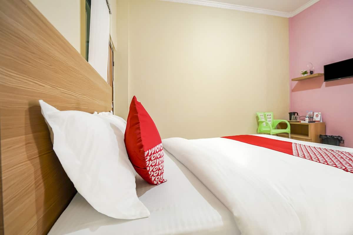 Ein ordentlich bezogenes Bett mit weißen und roten Kissen, ein Holz-Kopfschild und ein grüner Sessel in einem Zimmer mit beige- und rosa gestrichenen Wänden.