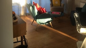 Un soggiorno con pavimento in legno, una poltrona verde, un divano beige e una sedia nera. Si notano delle librerie e una lampada sul lato destro.
