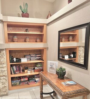 Un comptoir d’accueil avec une pancarte de bienvenue, des étagères en bois remplies de livres et de jeux de société, une plante en pot et un miroir.