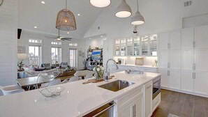 Une cuisine spacieuse avec des meubles blancs, un grand îlot central doté d’un évier, des luminaires suspendus et un ventilateur de plafond.