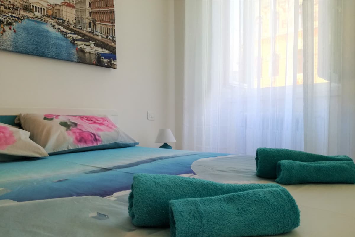 A hotel room with a bed, pillows, a lamp, and a painting of a cityscape.