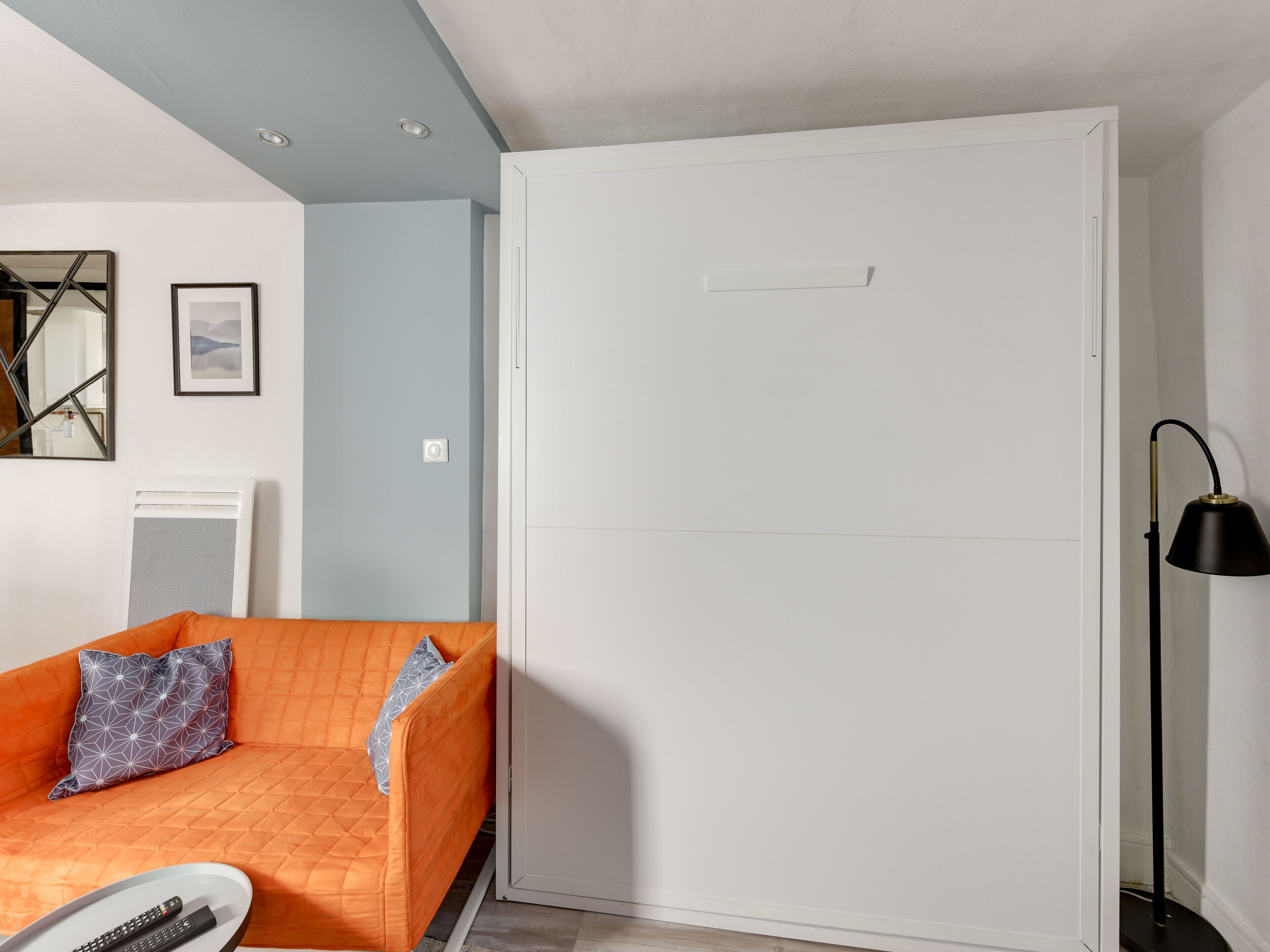 Ein Zimmer mit einem weißen Kleiderschrank, einem Bett mit orangefarbener Bettdecke, einer schwarzen Stehlampe und einer weißen Heizung.