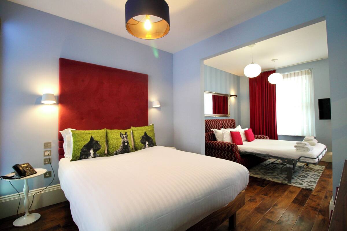 A hotel room with a large bed, a bedside table with a phone, a red accent wall, and a separate seating area with a sofa and red curtains.