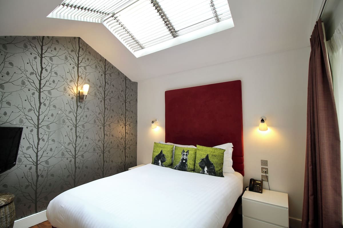 A hotel room with a bed, a red headboard, a nightstand with a phone, and a wall-mounted TV.