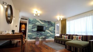 Un salon doté d’un plancher en bois, d’un ensemble de meubles de salon, d’un téléviseur fixé au mur et d’une grande fenêtre avec des rideaux.