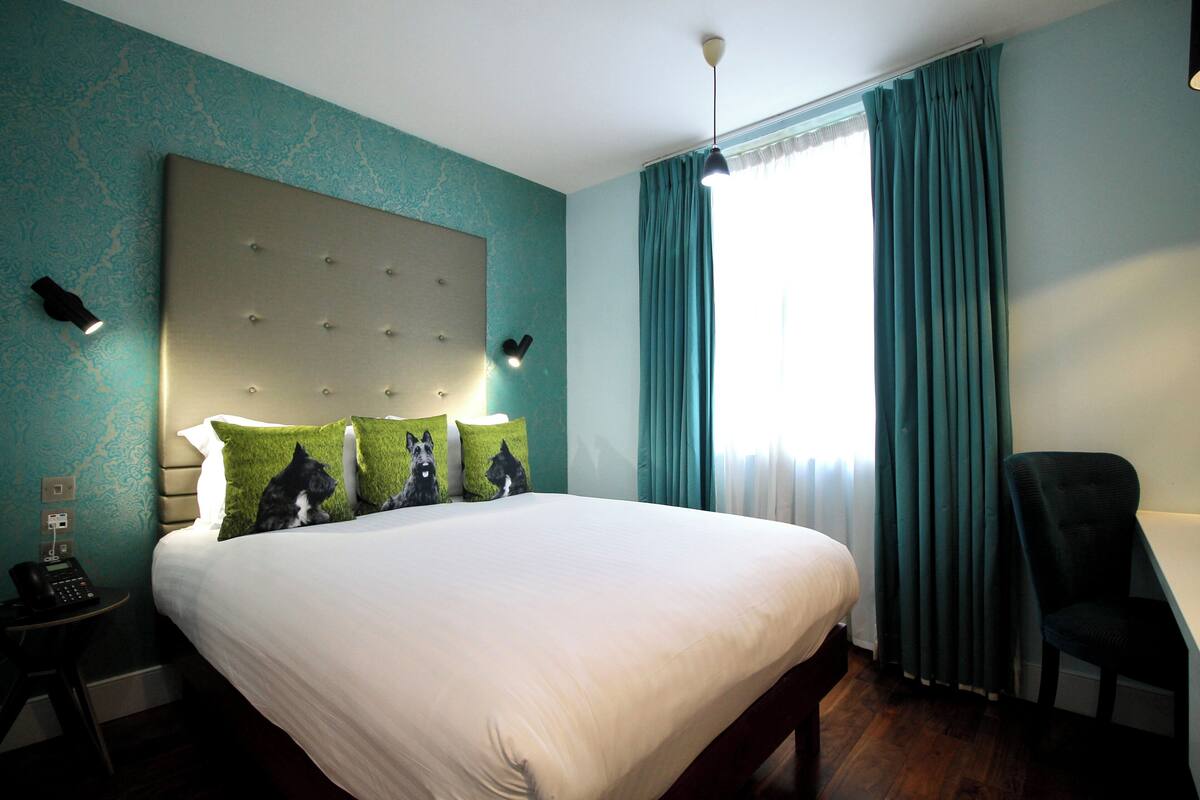 A hotel room with a large bed, a headboard with lights, a desk with a chair, and a window with curtains.