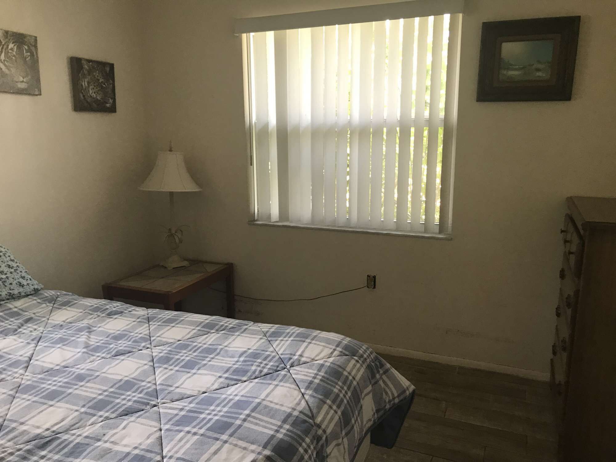 Une chambre à coucher comprenant un lit, une table de chevet, une lampe, une fenêtre avec des stores et une commode.