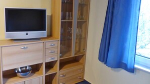 Una habitación con un televisor sobre un mueble de madera, una vitrina de cristal con estantes y una ventana con cortinas azules.