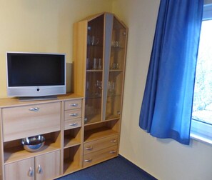 A room with a television on a wooden cabinet, a glass cabinet with shelves, and a window with blue curtains. - 2-room apartment on the upper floor with sunny balcony - Ferienhaus am Schmachter See (Binz)