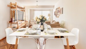 Un coin repas avec une table blanche dressée pour quatre personnes, entourée de chaises blanches. Un meuble TV en bois, sur lequel sont posées des bouteilles et un vase de fleurs, se trouve à gauche. Un canapé beige et une lampe sont visibles au fond.