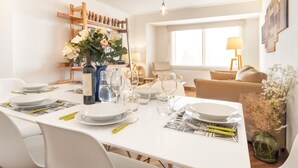 Un coin repas avec une table blanche dressée pour quatre personnes, entourée de chaises blanches. On aperçoit une étagère en bois avec des bouteilles et un vase de fleurs. Une fenêtre donnant sur l’extérieur est visible au fond.