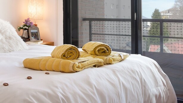 Une chambre d’hôtel avec un grand lit, un luminaire suspendu, une fenêtre donnant sur des immeubles et une petite table avec une lampe et des fleurs.