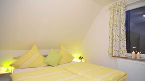 Ein Schlafzimmer mit Bett, Kissen, Nachttisch, Lampe und einem Fenster mit Vorhängen.