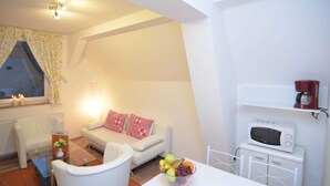 Ein kompakter Wohnraum mit einem weißen Sofa, einem kleinen Tisch mit Obstschale, einem weißen Stuhl, einem weißen Schrank mit Mikrowelle und einem Fenster mit Vorhängen.