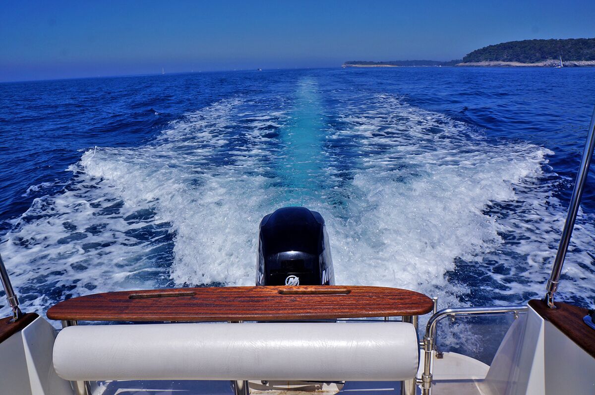 The bow of a boat with a wake in the water, a wooden deck, and a clear view of the sea and distant land.