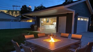 Une terrasse avec une table et des chaises carrées, un foyer extérieur et un espace couvert d’où l’on aperçoit une cuisine à l’arrière-plan.