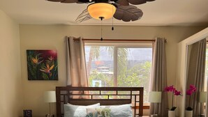 A bedroom with a large bed, a ceiling fan, a window with curtains, a nightstand with a lamp, and a painting on the wall. - 1 Bdrm Ocean View walk to Turtle Beach, close to Kona Village&Magic Sands Beach (Kailua)