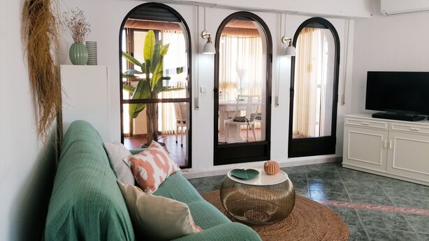 A living room with a green sofa, a round coffee table, and a television on a cabinet. There are two arched doorways leading to an outdoor area with a table and chairs. - A13 CONSULADO A 5º 3 (Calpe)