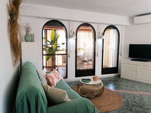 A living room with a green sofa, a round coffee table, and a television on a cabinet. There are two arched doorways leading to an outdoor area with a table and chairs. - A13 CONSULADO A 5º 3 (Calpe)