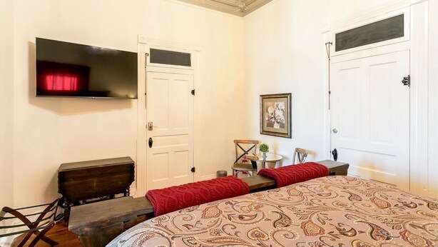 A bedroom with a large bed, a TV mounted on the wall, a door, and a small table with chairs.