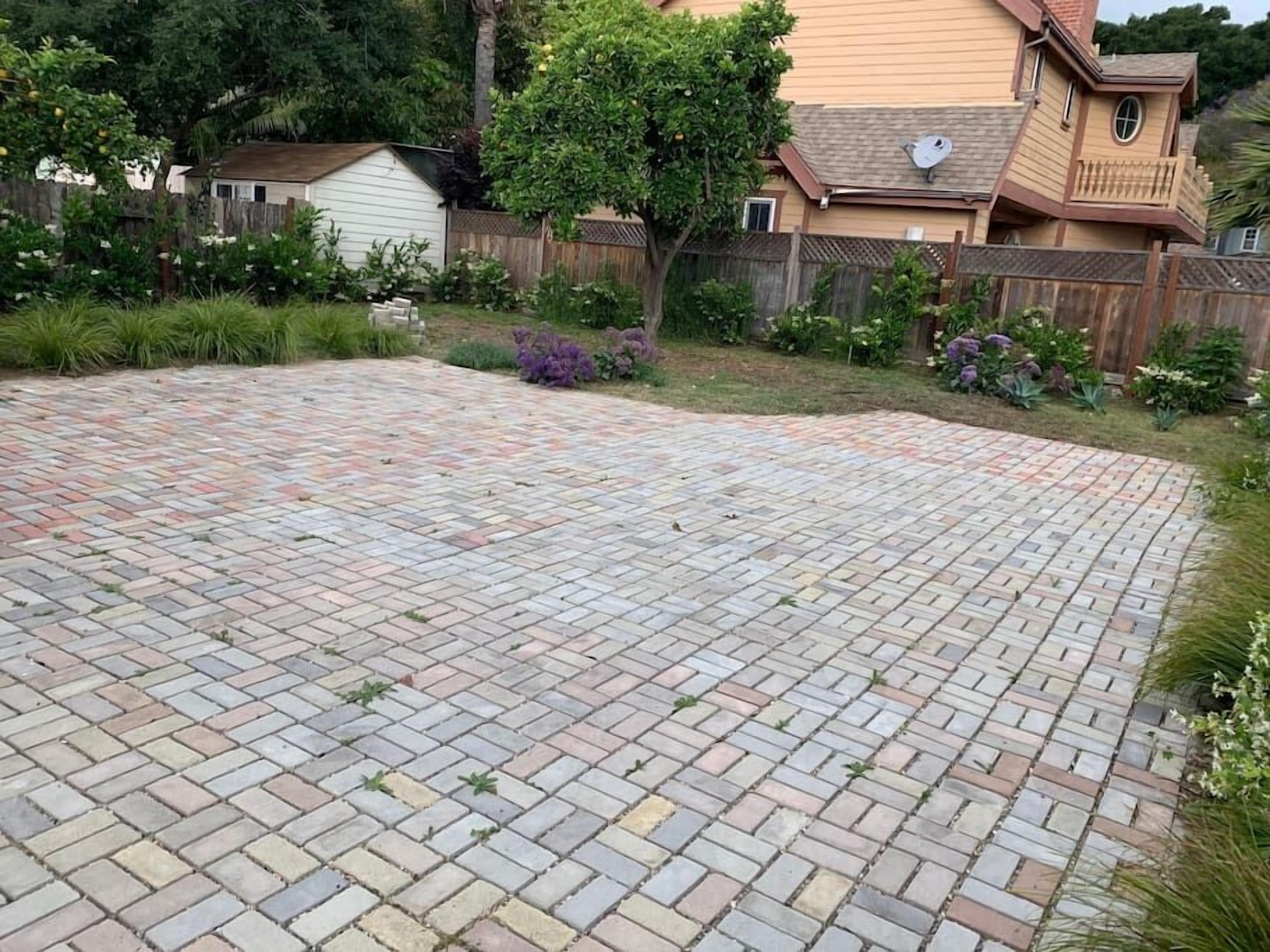 A paved backyard with a brick patio, surrounded by a wooden fence and various plants.