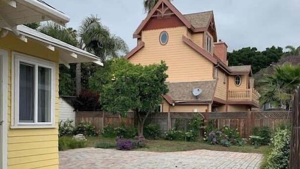 En have med en murstensbelagt terrasse, et gult hus og et stort hus i en unik, peachy farve med et særligt design.