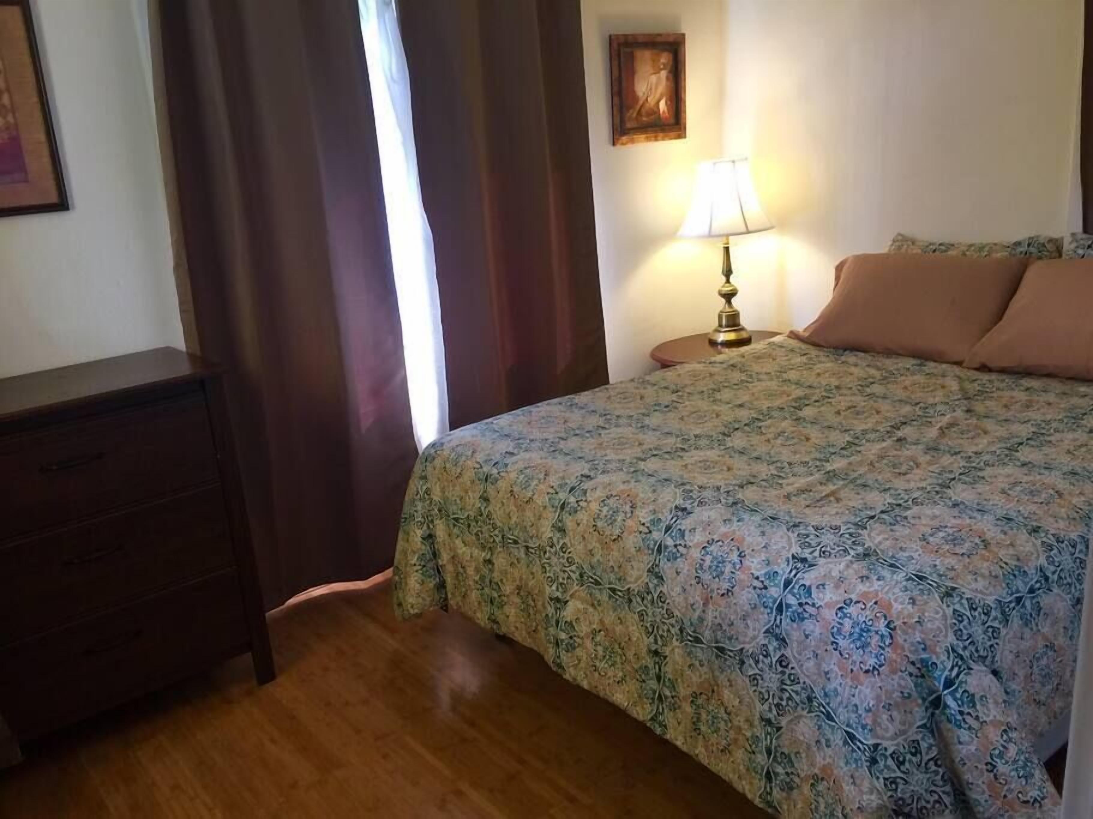 A hotel room with a wooden dresser, a bed with a floral bedspread, a nightstand with a lamp, and a painting on the wall.